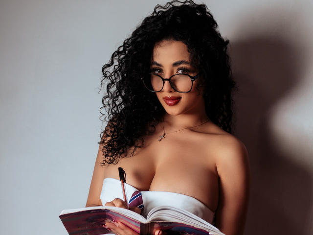 AuroraFerretti with curly black hair, wearing glasses and a strapless white top, reading a book, holding a pen, showing her tits, red lipstick, and a cross necklace.