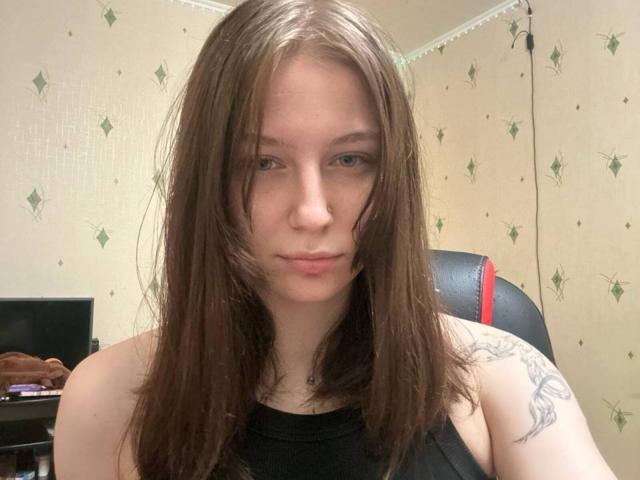 TonetteMasson with shoulder-length brown hair, slightly wet, looking at the camera with a neutral expression, black sleeveless top, tattoo on right shoulder, in a modestly decorated room.