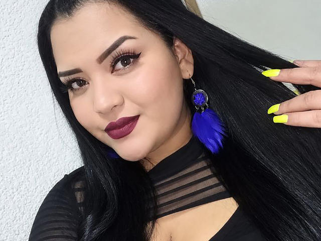 AlessiaLm con cabello negro largo, cejas oscuras, lápiz labial morado, uñas amarillas, aretes de plumas azules, blusa negra transparente, sujetando el cabello, sonriendo, piel clara, linda, sexy..