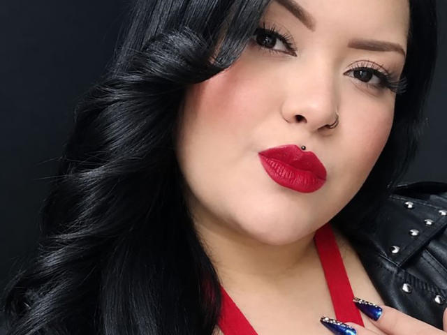 AlessiaLm hat schwarzes, welliges Haar, roten Lippenstift, einen Nasenring und eine schwarze Jacke mit Nieten, kombiniert mit einem roten Top mit Halter, und blickt mit leicht zusammengepressten Lippen in die Kamera..