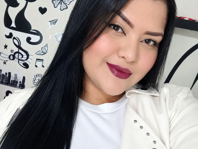 AlessiaLm mit langen, schwarzen Haaren, dunklen Augenbrauen, einem Nasenring und glänzendem, violettem Lippenstift, trägt eine weiße Jacke über einem weißen Hemd. Sie lächelt leicht..