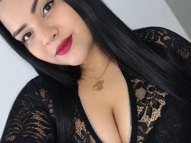 AlessiaLm con una blusa negra de encaje, que deja ver parte del escote, usando lápiz labial rojo, un collar dorado y cabello negro largo, sonriendo a la cámara. Se ven los senos a través del encaje..