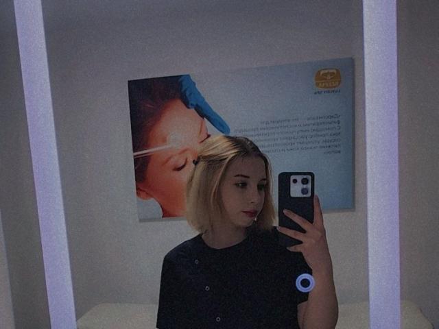 LavonneAddy with blonde bob, dark shirt, holding phone, mirror selfie, blue-gloved woman's face in background, serious expression, slight red lipstick, white wall, visible ring light.