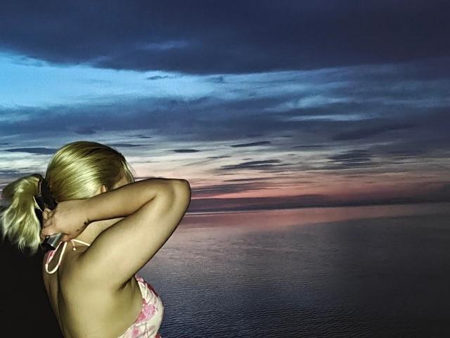 LavonneAddy with blonde hair in a ponytail, tying a pink floral bikini top, showing her back and bare ass, against a dark, cloudy sunset over a calm ocean.