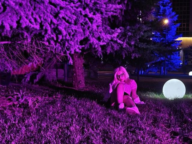 LavonneAddy, blonde, sitting on grass, wearing glasses and a sparkly top, under purple-lit tree, with glowing orb to the right, looking at camera.