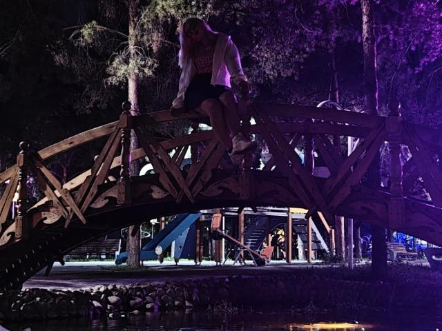 LavonneAddy sits on a wooden bridge at night, wearing a white shirt and black shorts, with purple and blue lights illuminating the scene and a dark background.
