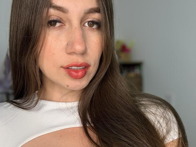 Maittee has long brown hair, brown eyes, and red lipstick. She's wearing a white top, showing her tits slightly. Her nose has a small mole. She's looking at the camera.