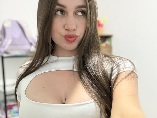 Maittee with long brown hair, white cut-out top, showing her big tits, pouty pink lips, and looking to the side, in a selfie. Her skin is fair and she's cute.