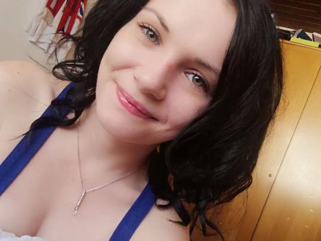 Elliefucku30-hot heeft een lichte huid, zwart golvend haar, groene ogen en een lichte glimlach. Ze draagt een blauwe top met een witte kantrand, waardoor haar borsten zichtbaar zijn..