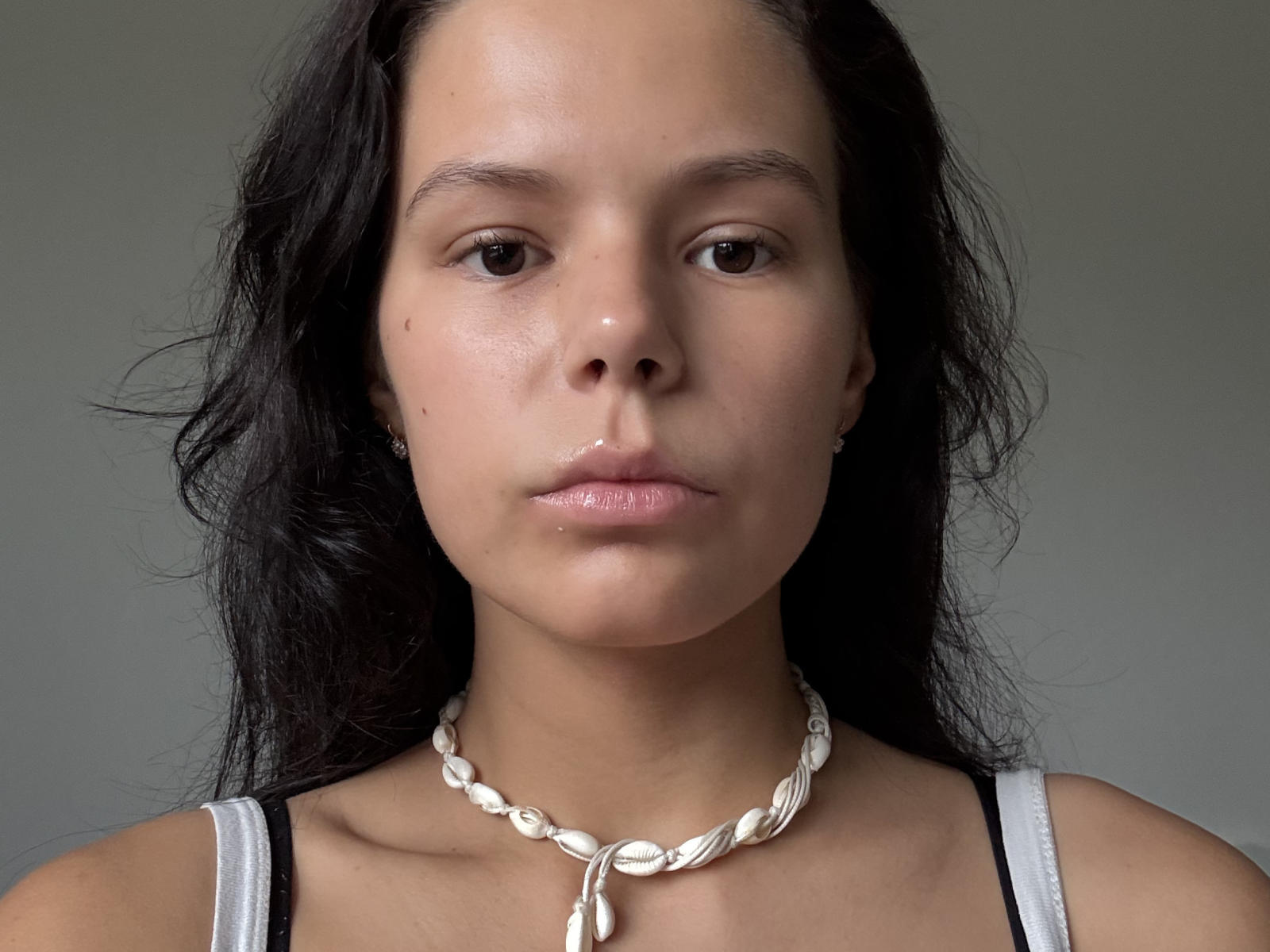 ClarenceGarin has light brown skin, dark wavy hair, small dark eyes, and a neutral expression. She's wearing a white seashell necklace and a white tank top.