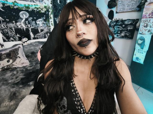 DollyBones has dark, wavy hair, black lipstick, and a nose ring. She's wearing a black, spiked choker and a low-cut, black top. Her tits are partially visible.