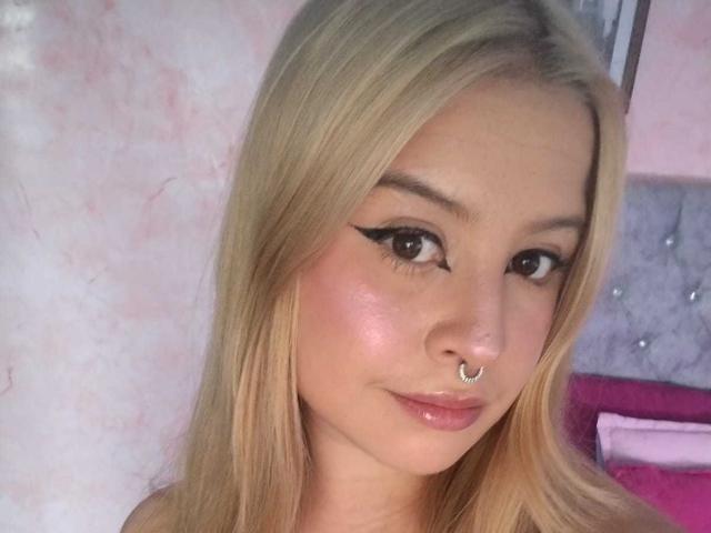 MelaniBecker has blonde hair, a nose ring, black eyeliner, and glossy pink lips. Her skin is shiny, and she's looking at the camera with a slight smile.