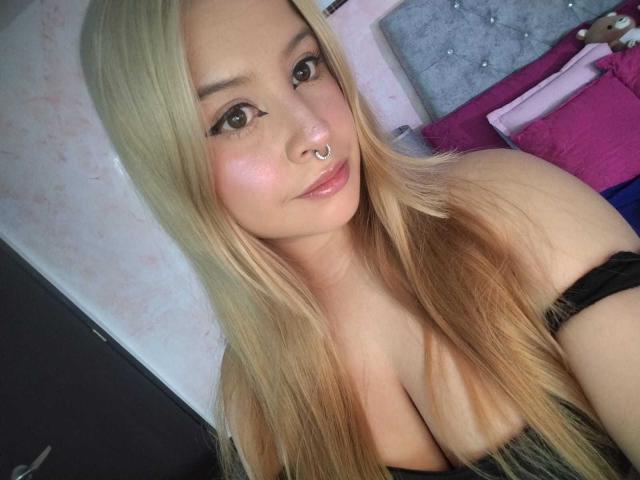 MelaniBecker with long blonde hair, nose ring, and star-shaped eyeliner, showing her cleavage in a black strapless top, smiling at the camera.