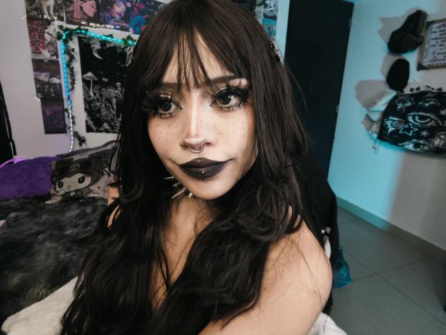 DollyBones with long black hair, dark lipstick, heavy eyeliner, and a nose ring, sitting in a room with a black and white patterned wall. Boobs partially visible.