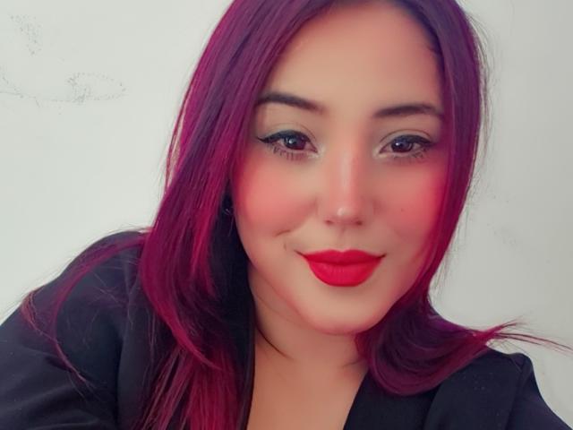 Thalia Sorner, con cabello rosa intenso, lápiz labial rojo, delineador de ojos negro y una sonrisa encantadora, lleva una blusa negra. Su trasero no es visible, solo su rostro..