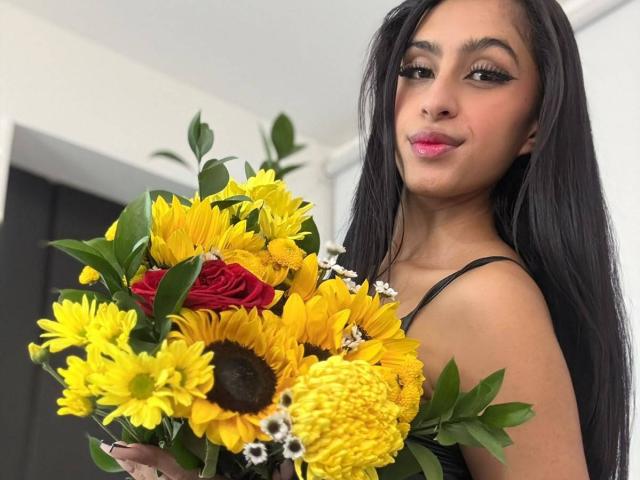 LilithDesire with long black hair, pink lipstick, holding a sunflower bouquet, featuring yellow flowers, red roses, and small white flowers. Her tits are slightly visible under a black top.
