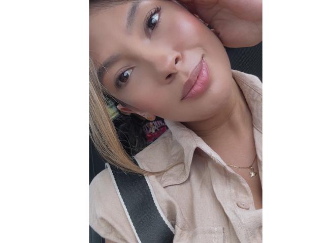 KendraMiler has a cute smile, subtle makeup, and is wearing a beige shirt with a small necklace. She's in a car seat, with her hand near her head.