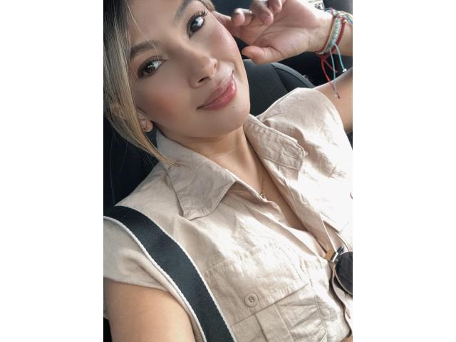 KendraMiler with cute, smiling face, light skin, blonde hair, wearing beige shirt, black straps, colorful bracelets, subtle makeup, slightly open shirt, showing a hint of cleavage.