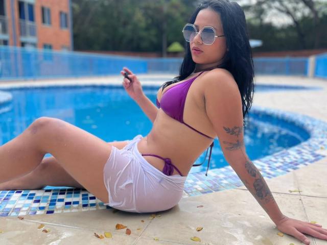 AlanaLuxe with dark hair, wearing a purple bikini top and white wet shorts, sitting by a blue pool, sporting sunglasses, tattoos on her arm, and holding a black object.
