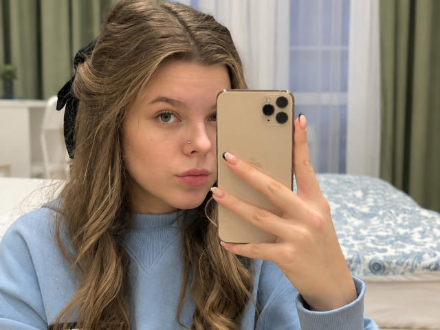 RhonaBells with wavy brown hair, light blue sweater, and black bow, taking a mirror selfie with a gold iPhone, pouty lips, nose ring, and white nails.