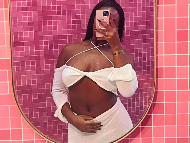 VenusWilly with dark skin, wearing a white off-shoulder dress, showing her tits and pussy under the dress, taking a mirror selfie with a pink phone.