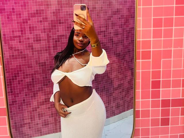 VenusWilly with dark skin, long black hair, white off-shoulder top, and white skirt, showing her tits and ass, holding a pink phone, standing in front of a pink tiled wall.