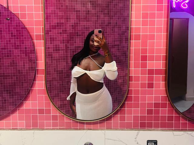 VenusWilly, avec une peau foncée, de longs cheveux noirs, portant un haut blanc à une épaule et une jupe, montrant ses seins et ses fesses dans un selfie pris devant un miroir, tenant un téléphone rose..