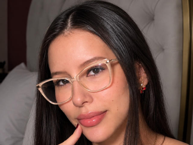 AlisJenner has long dark hair, wearing clear glasses, pink glossy lips, and red earrings, with her finger touching her chin, looking cute and sexy.