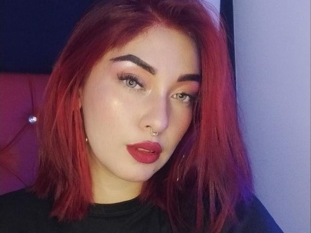 BriielaEva, com cabelo ruivo, piercing no nariz e batom vermelho, olhando para a câmera com um leve franzido, vestindo uma camisa preta. Ela tem olhos verdes e um rosto brilhante..