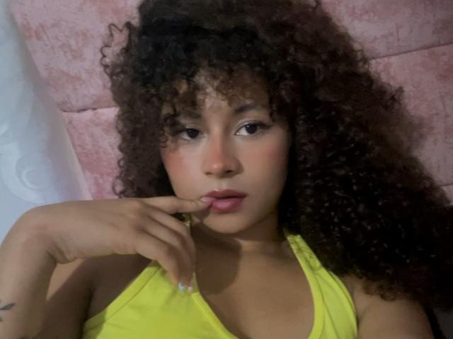 SofiaPassionne, con cabello castaño rizado, vestida con una blusa amarilla, mordiéndose el dedo, mirando a la cámara, con un pequeño tatuaje en la mano izquierda. Su vulva no es visible..