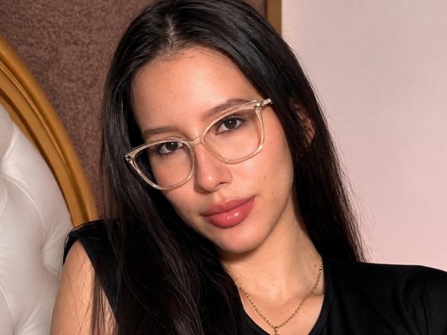 AlisJenner has long black hair, wears clear glasses, and a black top, with pinkish lips and a gold necklace, looking straight at the camera.