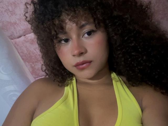 SofiaPassionne has curly brown hair, brown eyes, and a cute face with pink cheeks. She's wearing a yellow halter top, showing her small tits. Lying down, looking at camera.