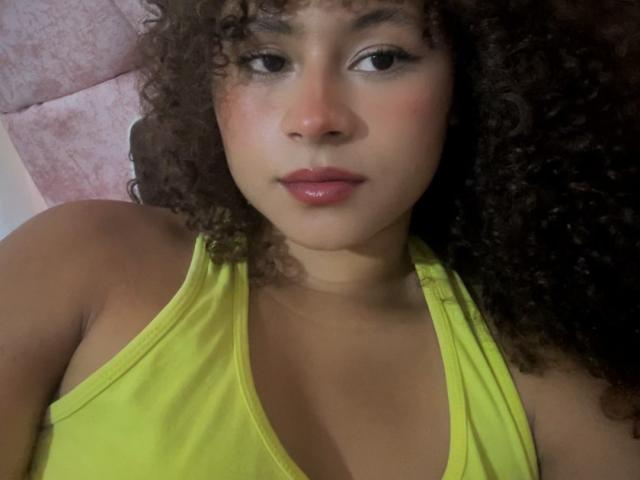 SofiaPassionne with curly brown hair, wearing a yellow tank top, showing her bare shoulders and small tits, has pink lips and a slight blush on her brown skin.