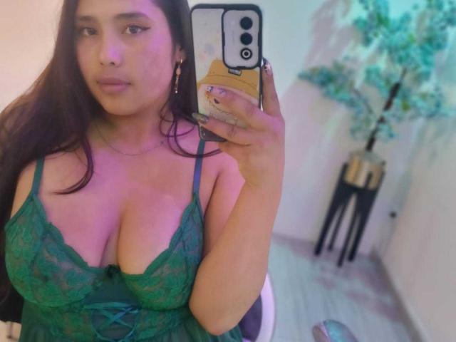 GemmaBellini with large tits in a green lace babydoll, holding a phone in a mirror selfie, has long brown hair, pinkish lighting, and a potted plant in the background.