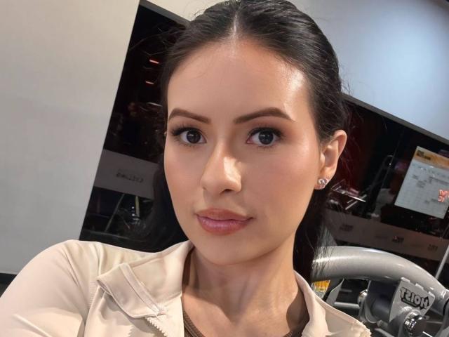 HannaRobertss with dark hair, blue eyes, and pink lipstick, wearing a beige jacket, showing off her smooth, shiny face and small, diamond-studded earrings, smiling slightly.