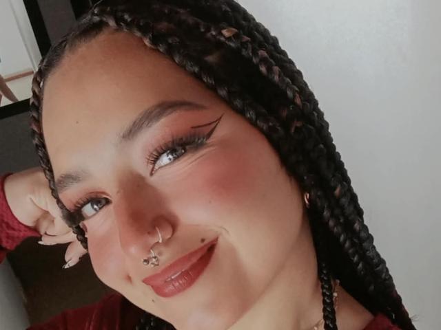 TatianaMoora with braided hair, nose ring, lip piercing, and red lipstick, smiling at the camera. She has pink eye makeup and long eyelashes. Boobs not visible.