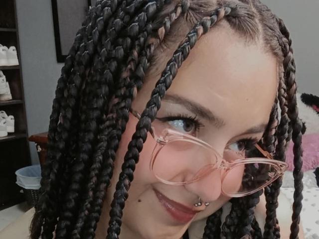 TatianaMoora, avec ses cheveux noirs tressés, ses lunettes roses, son piercing au nez et son rouge à lèvres rouge, sourit à l'appareil photo. Ses seins sont partiellement visibles, et elle affiche un sourire discret..