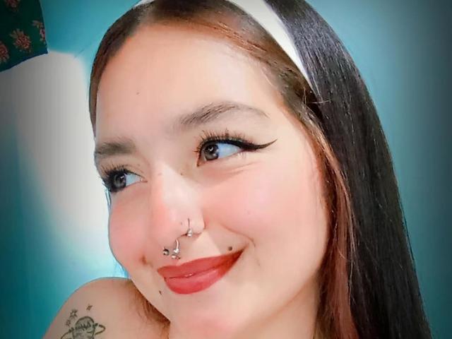 TatianaMoora hat ein hübsches Gesicht mit rotem Lippenstift, schwarzem Eyeliner, Nasenpiercings, einem Septum-Ring und einem weißen Stirnband. Ihre braunen Haare und ein Tattoo sind sichtbar..