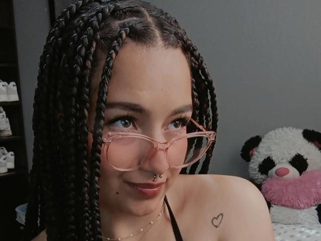 TatianaMoora, com cabelo preto trançado, óculos rosa, piercing no nariz e tatuagem de coração no ombro, sorrindo para a câmera. Seios parcialmente visíveis, vestindo um top preto de alças..