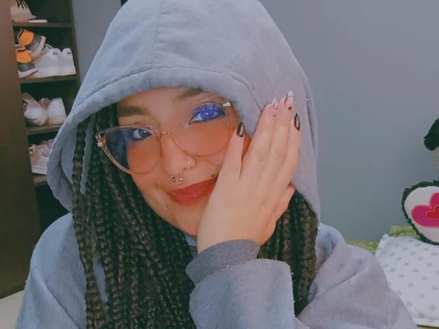 TatianaMoora, con cabello negro trenzado, ojos azules, gafas, aro en la nariz y aro en el tabique, y una sudadera gris, con la mano en la mejilla, sonriendo. Uñas nude transparentes, sudadera gris, aspecto casual..