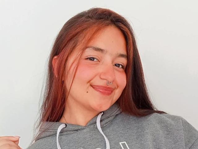 TatianaMoora has brown hair, a nose ring, and a smile, wearing a gray hoodie with white drawstrings. Her tits are small, and she has a cute mole on her chin.