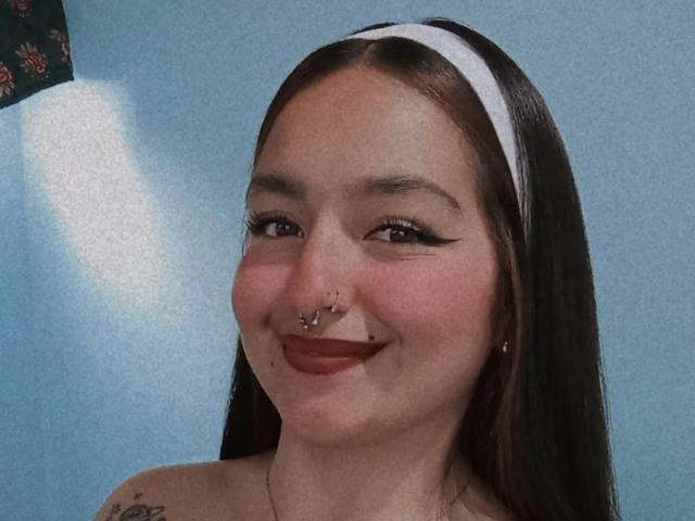 TatianaMoora has brown hair, a white headband, nose piercings, a lip piercing, and winged eyeliner, smiling with red lipstick, and a tattoo on her shoulder.