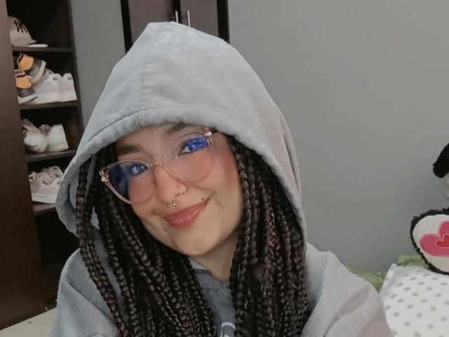 Tatiana Moora, com cabelo preto trançado, vestindo um moletom cinza e óculos com lentes azuis, sorrindo com um piercing no nariz e olhos azuis. Seus seios não estão visíveis..