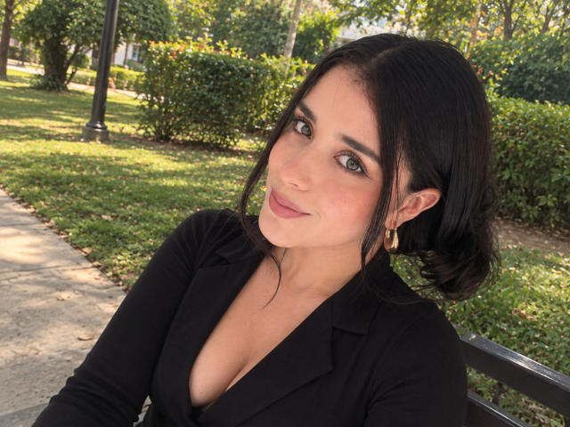 VickyMegan with dark hair, green eyes, and gold earrings, wearing a black top with a deep cleavage, sitting outdoors, smiling softly at the camera.