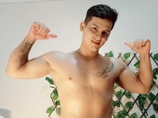 Jeff Tayler ohne Hemd, mit kurzen schwarzen Haaren, die Arme anspannt, Tätowierungen am linken Arm und an der Brust zeigt, lächelnd, mittelgroßer Statur, helle Hautfarbe, steht vor einer grünen Wand mit Blättern..
