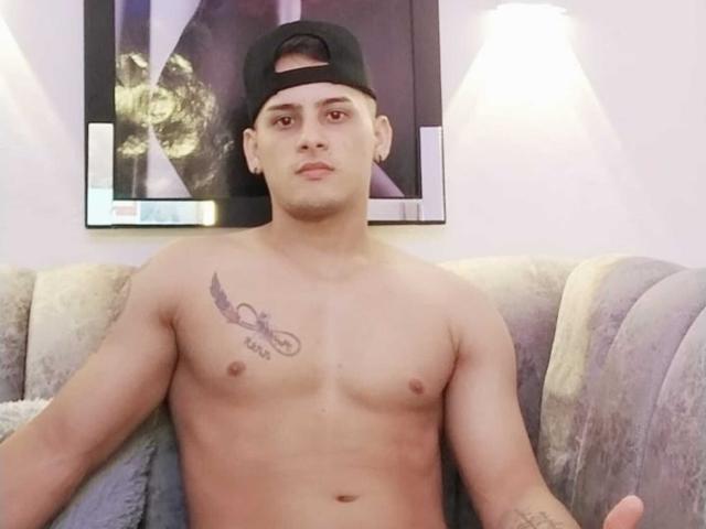JeffTaylers sans chemise, avec une peau brun clair, des cheveux bruns courts et une casquette noire, assis sur un canapé gris, montrant un tatouage sur sa poitrine gauche, regardant la caméra..