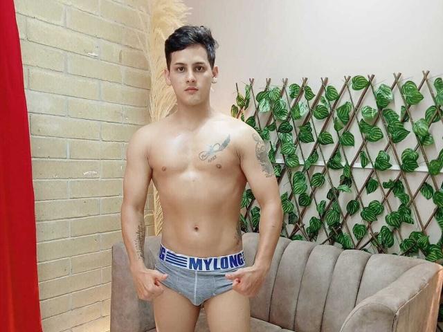 Jeff Taylers, ohne Hemd, mit Tattoos, kurzes schwarzes Haar, stehend, trägt graue NYLONG Boxershorts, Hände an der Hüfte, vor einer beige gefliesten Wand und einer Wand mit grünem Blattmuster..