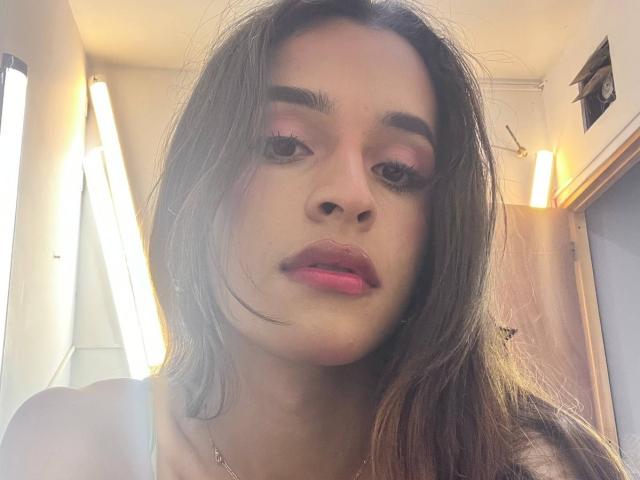 AmyLevins, con lápiz labial rosa, cabello castaño y maquillaje de ojos discreto, mirando a la cámara con una ligera mueca, vestida con una blusa blanca y un collar de plata..