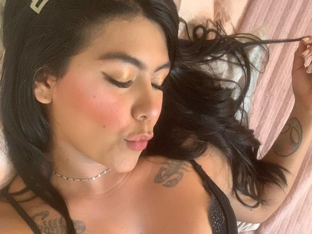CatalinaReiyna with black hair, closed eyes, pink blush, and tattoos, pouting lips, wearing a black lace bra, lying on a pink pillow, holding a strand of hair.