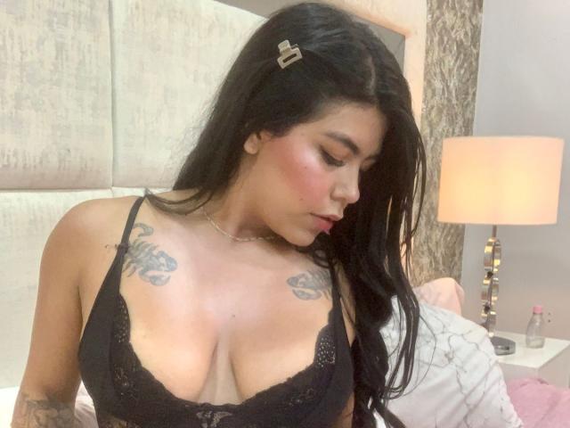 CatalinaReiyna with black lace bra, showing her big tits, tattoos on her chest, long black hair, looking down, red blush on her cheeks, in a bedroom.